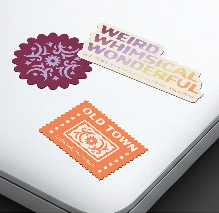 Laptop decorated with Old Town branded stickers. One reads, "Old Town, Lansing Michigan" featuring the floral orange brick icon of the logo. Another reads, "Weird, Whimsical, Wonderful."