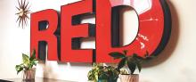 A large red sign in block text reads "RED" on a white wall, with plants. 